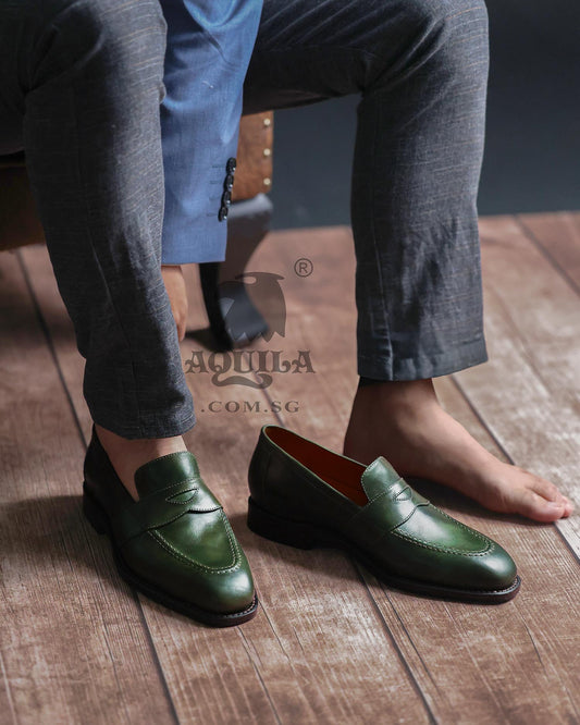 Green leather loafers worn with gray pants on a wooden floor, Aquila brand visible.